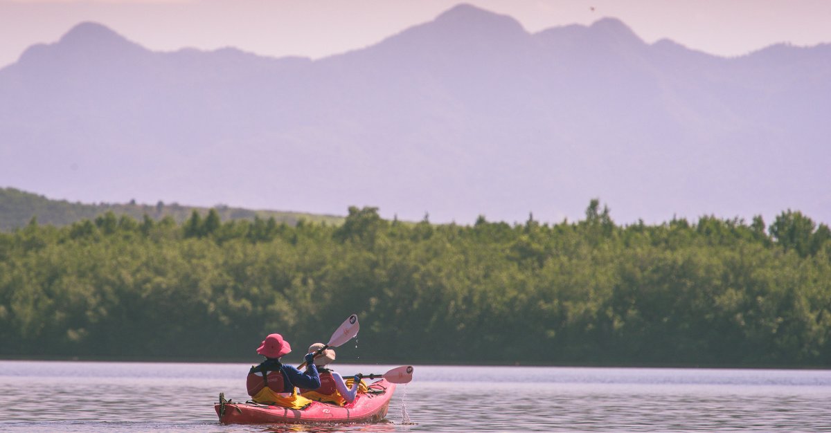 Two people in sun hats paddling a red tandem sea kayak on Caribbean waters with Cuban mountains in front of them.