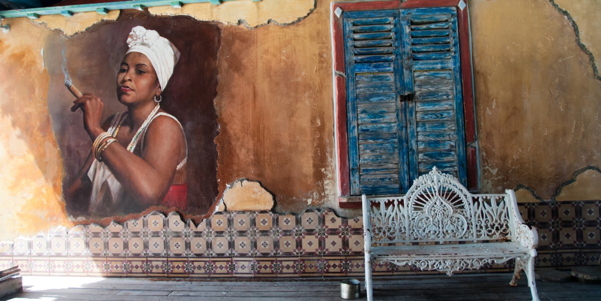 Outside of an old building in Cuba with a painting of a Cuban woman smoking a cigar and a white bench in front of the mural