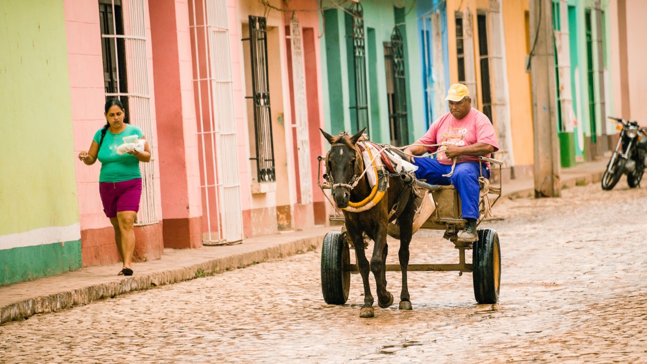 Person in a horse and carriage ride on a cobblestone road while a woman walks on the sidewalk among colorful buildings