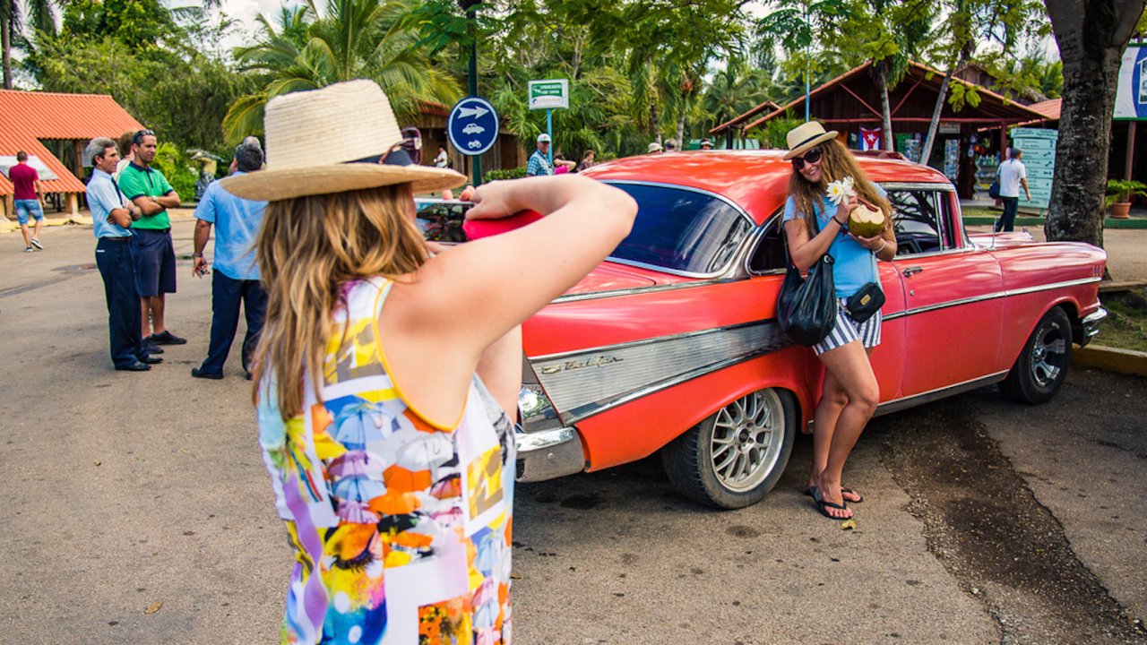 A woman taking a picture of another woman posing in front of a classic red Cuban car.