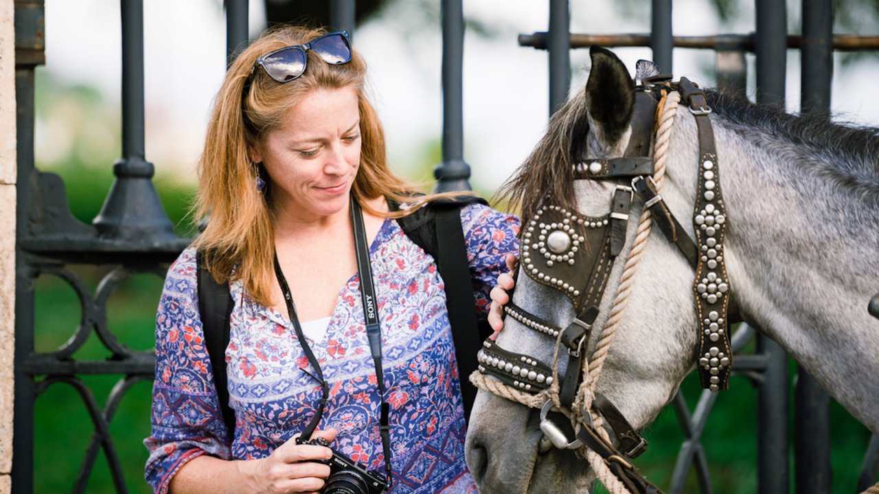 A woman standing petting a horse with a camera around her neck.