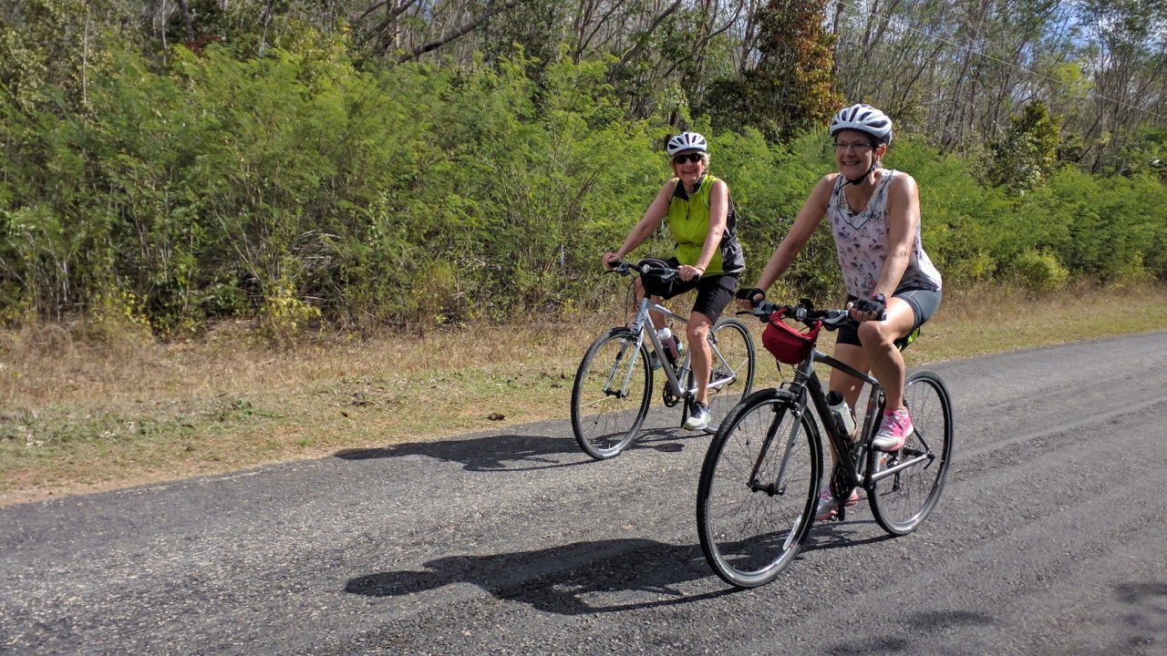 Two women enjoy a sunny day riding road bikes along a quiet highway surrounded by nature during a cycle Cuba tour designed for active travelers.